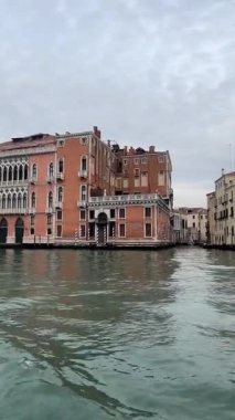 Büyük ve karmaşık bir şekilde tasarlanmış pembe Venedik sarayı Büyük Kanal boyunca belirgin bir şekilde duruyor ve bulutlu bir gökyüzünün altındaki sakin su üzerindeki detaylı mimari unsurlar ve yansımaları sergiliyor..