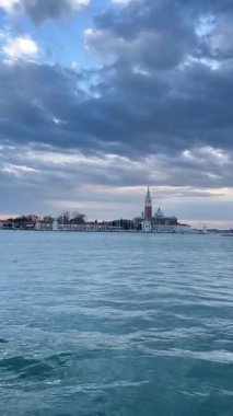 Venedik 'teki San Giorgio Maggiore Adası' nın panoramik manzarası, ikonik kilisesi ve çan kulesi, dramatik bulutlu gökyüzüne ve alacakaranlıkta sakin lagün sularına karşı silueti..