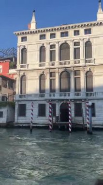 Klasik mimarisi ve koyu renk kepenkleri olan büyük beyaz Venedik sarayı güneş ışığıyla aydınlatılmış, ön planda kırmızı ve beyaz demirleme direkleri bulunan bir kanal boyunca zarifçe duruyor..