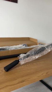 Bir kişinin eli, bir barfiks çubuğunun siyah metal bileşenini tutuyor, ana ambalaj kutusu ve diğer parçaları tahta bir yüzey üzerinde görünür durumda..