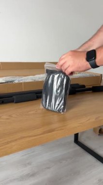 Bir kişinin elleri, içinde yeni bir barfiks çubuğu bulunan açık karton kutudan plastik aksesuar ve siyah bir kayış çıkarırken görülüyor..