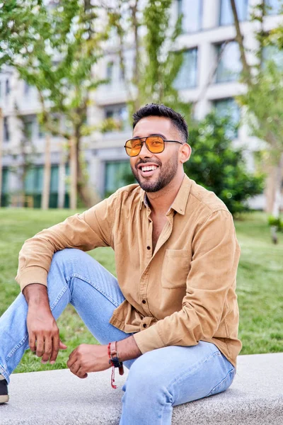 Young happy Hispanic man thinking while sitting at park. latin man wearing sunglasses