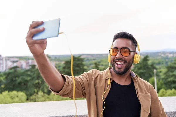 latin man with cell phone in hand making a video call. latin man with beard and sunglasses wearing yellow headphones.