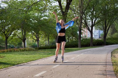 Parkta patenci kız paten kayıyor, kollarını kaldırıyor.