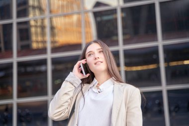Neşeli, genç, çekici, esmer kadın cep telefonundan gülümsüyor. Modern ofis. Yüksek binaların arka planı.