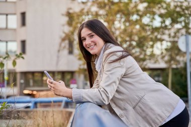 Korkuluklara yaslanmış elinde cep telefonuyla kameraya bakan genç bir öğrenci.