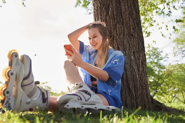 Woman skating in park. Girl skating sitting on grass relaxing with inline skates while chatting on smart phone. Caucasian woman in outdoor activities.