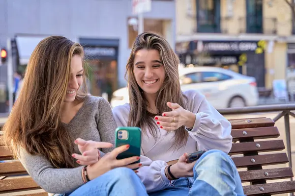 two women laughing while looking at their cell phones sitting on a city bench. couple of women addicted to social networks.