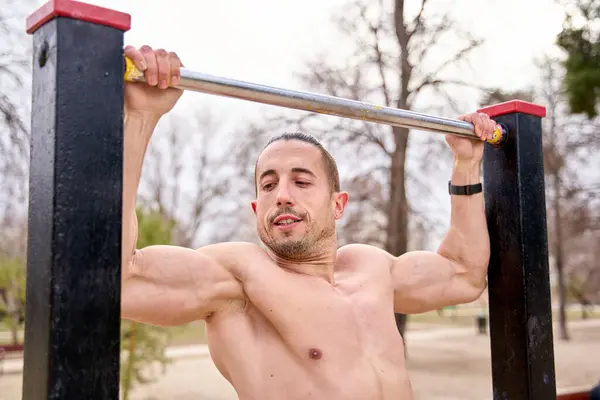 Fitness Man Doing Pull-Ups At The Bars In A City Park. Front view of a strong man doing pull-ups in the sports park at sunset during the summer.