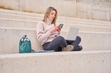 Genç mutlu sarışın kız öğrenci dizüstü bilgisayar kullanıyor ve cep telefonu öğreniyor üniversite parkında oturuyor, çevrimiçi dijital uzaktan eğitim görüyor ya da dışarıda öğrenim görüyor..