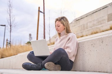 Üniversite kampüsündeki bankta oturan genç bir öğrenci dizüstü bilgisayarında çalışıyor.