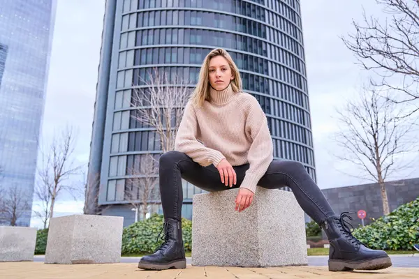full-length photograph of a blonde woman in casual clothes sitting on a stone bench in the city while looking at the camera.