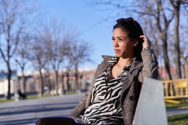 Genç Latin kız şehirde bir bankta oturuyor ve bekliyor ve dinleniyor.