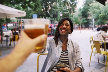 Gülümseyen bir adam restoranda oturuyor, şık bir kıyafet giyiyor, telefon kullanıyor ve bira içiyor..