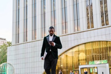 Siyah işadamı elinde takım elbise ve dizüstü bilgisayarla ofisten çıkıyor.