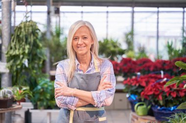 Orta yaşlı kadın küçük çiçekçi dükkânı sahibi dikilip fotokopi alanı olan kameraya bakıyor.