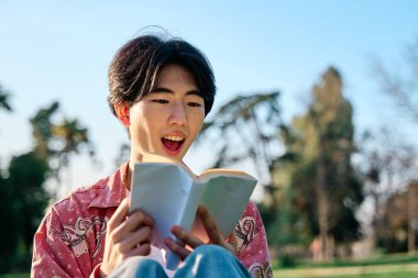 Genç Asyalı bir adam oturuyor ve yüzünde şaşırmış bir ifadeyle kitap okuyor..