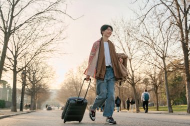 Genç Asyalı bir adam bir bavul taşıyarak şehirde yürüyor. Gülümseyen ve kendine güvenen adam.