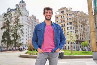 Şehirde sıradan kıyafetler, kot pantolon ve gömlek giymiş bir adamın resmi. Kameraya bakıyor.