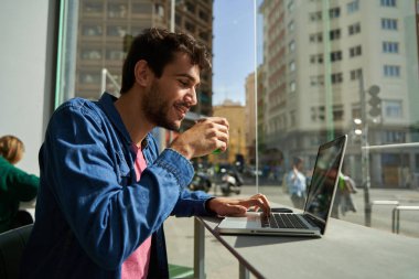 Yakışıklı sakallı hippi, kafede kahve masasında dizüstü bilgisayarı kullanıyor ve ona bakıyor. İletişim ve teknoloji kavramı