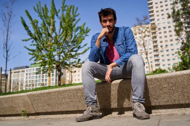 Şehirdeki taş bir bankta oturan genç adam kameraya bakıyor.
