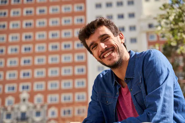 Mavi gömlekli bir adam gülümsüyor ve bankta oturuyor. Arkasındaki bina kırmızı ve beyaz.