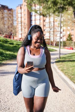 Genç bir Afro-Amerikan kadının akıllı telefonunu tutarken kulaklıkla müzik dinlemesinin dikey portresi. Kıvrımlı bir kadın spor salonuna giderken gülümsüyor..