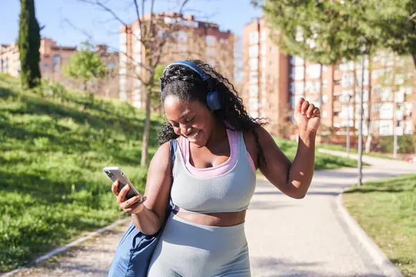 Afro-Amerikan kadın portresi spor kıyafetleriyle yürürken akıllı telefonuyla kulaklıkla müzik dinliyor..