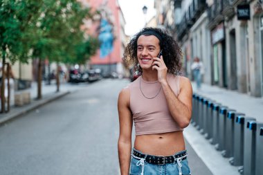 Uzun kıvırcık saçlı genç eşcinsel adam akıllı telefondan konuşurken kasabada dolanıyor.