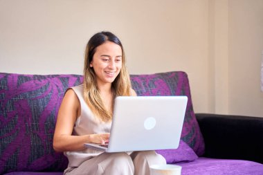 Dizüstü bilgisayar kullanan gülümseyen genç bir kadın, evdeki kanepede oturuyor, güzel bir kız alışveriş yapıyor ya da sosyal ağda sohbet ediyor, eğleniyor, film izliyor, bilgisayar projesinde serbest çalışıyor.
