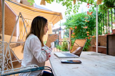 Evdeki terasta açık bir masada dizüstü bilgisayarda çalışan genç bir kadının yan görüntüsü. Elinde bir fincan çay ya da kahve tutuyordu..