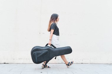 Beyaz kadın kaldırımda gitar çantası taşıyor.