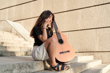 Genç, esmer, Latin bir kadın gitarla oturmuş akıllı telefonunu kullanıyor.