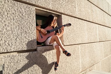 Duvar teneffüsünde oturan bir kadının tam görüntüsü. Metin için kopyalama alanı ile gitar çalıyor. minimalist fotoğraf.