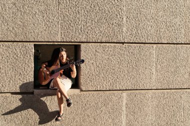 Duvar teneffüsünde oturan bir kadının tam görüntüsü. Metin için kopyalama alanı ile gitar çalıyor. minimalist fotoğraf.