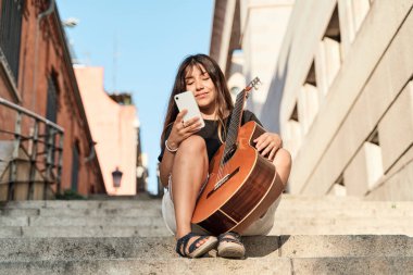 Genç, esmer, Latin bir kadın gitarla oturmuş akıllı telefonunu kullanıyor.