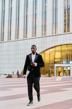 Siyah işadamı elinde takım elbise ve dizüstü bilgisayarla ofisten çıkıyor.
