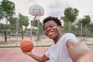 Genç Afrikalı Amerikalı bir adam elinde topu tutarken akıllı telefonuyla bir basketbol sahasında selfie çekiyor.