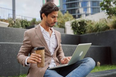 Genç ve zarif bir adam sıcak bir içecek ya da kahve içerken bilgisayarını kullanarak internette çalışıyor.