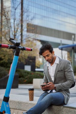 Genç işadamı, arka planda ofis binaları ve yanında elektrikli scooterıyla taş bir bankta oturan akıllı telefonunu kullanıyor.