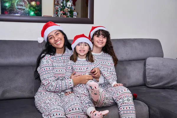 Noel pijamaları ve Noel Baba şapkaları giymiş kız kardeşler kanepede oturuyor.