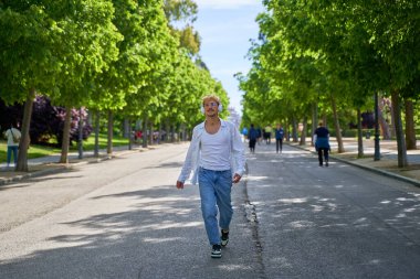 Beyaz gömlekli ve kot pantolonlu bir adam arka planda ağaçlarla caddede yürüyor. Kulaklık takıyor ve müzik dinliyor.