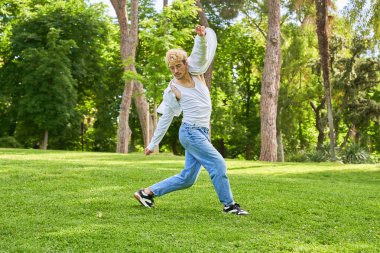 Kot pantolonlu ve beyaz tişörtlü genç bir adam parkın ortasında modern dans ediyor.