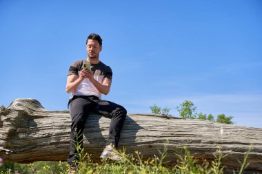 Adam akıllı telefonunu kullanarak devrilmiş bir ağaçta oturuyor.