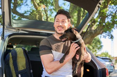 Köpeğini okşayan bir adam arabanın bagajında oturuyor. Seyahatteler ve gülümsüyorlar..