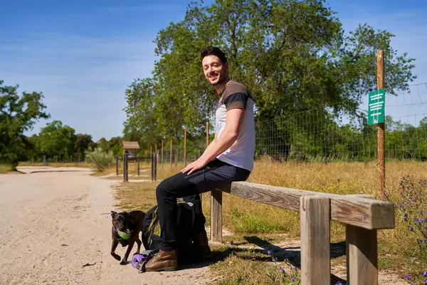 Bir adam köpeğiyle bankta oturuyor. Köpek mor bir tasma takıyor. Adam gülümsüyor ve evcil hayvanıyla eğleniyor.