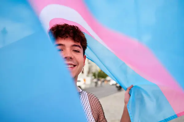 Bir adam gülümsüyor ve elinde bir trans bayrak tutuyor. Arkasında bayrak var ve beyaz bir gömlek giyiyor.