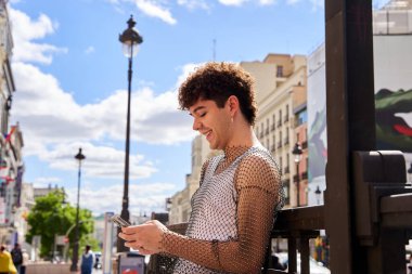 Kıvırcık saçlı bir adam bankta oturuyor ve telefonuna bakıyor..