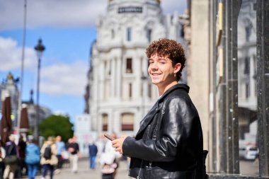 A young gay man in a leather jacket is smiling and holding a cell phone. He is standing in front of a building with a crowd of people around him clipart