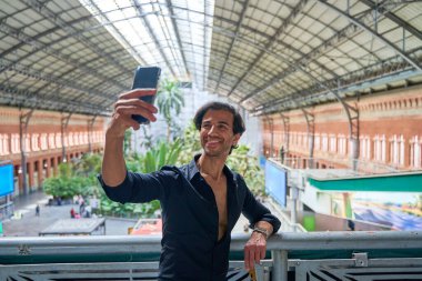 Siyah tişörtlü Hintli bir adam tren istasyonunda selfie çekiyor. Arka planda yüksek tavan ve yeşillik var..